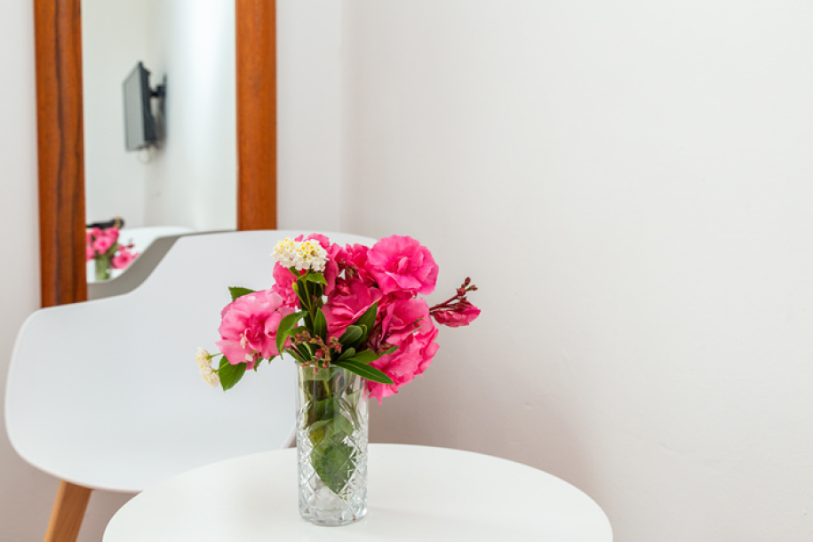 Vaso di fiori rosa su tavolo bianco con specchio e sedia sullo sfondo.